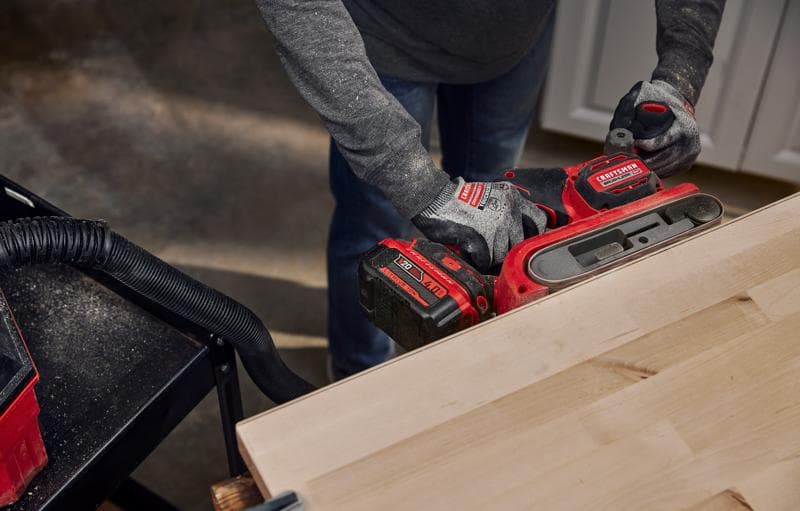 Photo d'une personne utilisant une ponceuse à bande sans fil CRAFTSMAN, SKU CMCW223B, ponçant une surface en bois dans un atelier.