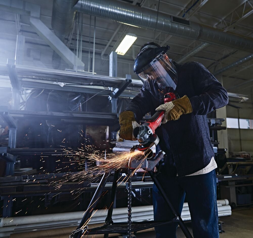 Photo of worker using CRAFTSMAN angle grinder CMCG450B, producing sparks while cutting metal pipes in an industrial workshop.