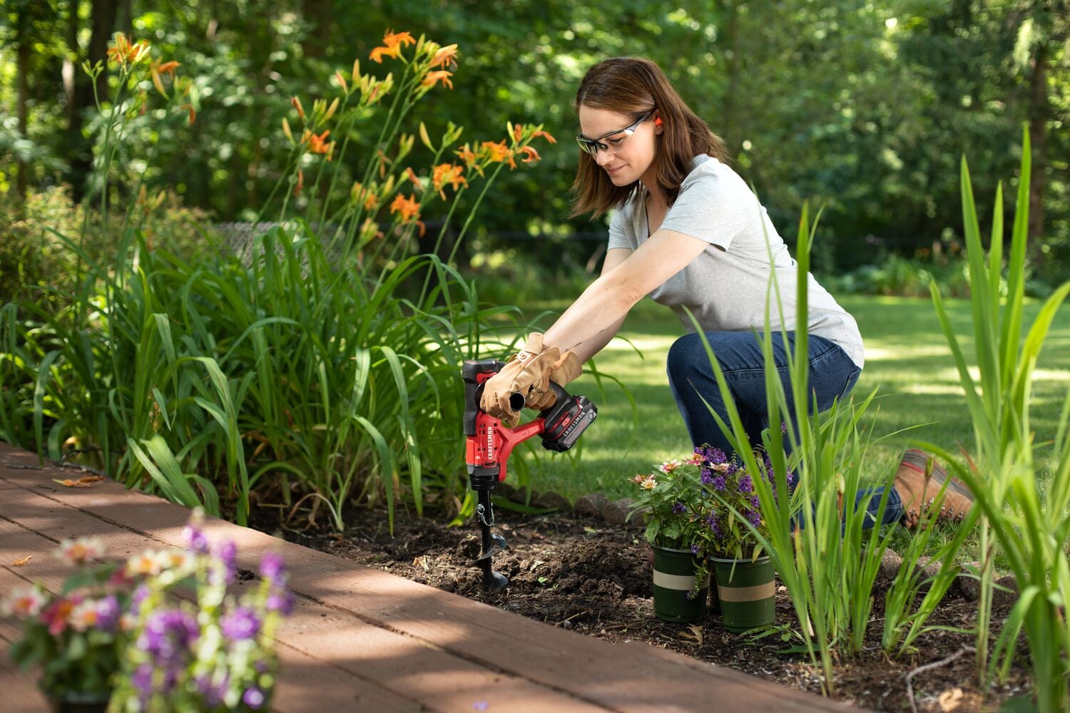 Woman using CRAFTSMAN CMCA320C1 cordless auger in garden bed, surrounded by flowers and plants.