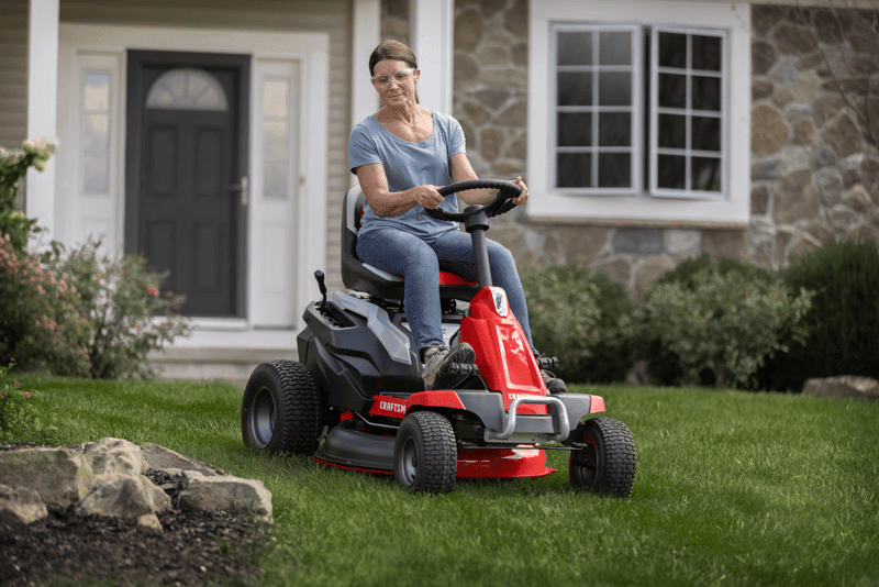 CRAFTSMAN Battery-Powered Compact Mower cutting grass around flowerbed with house in background.