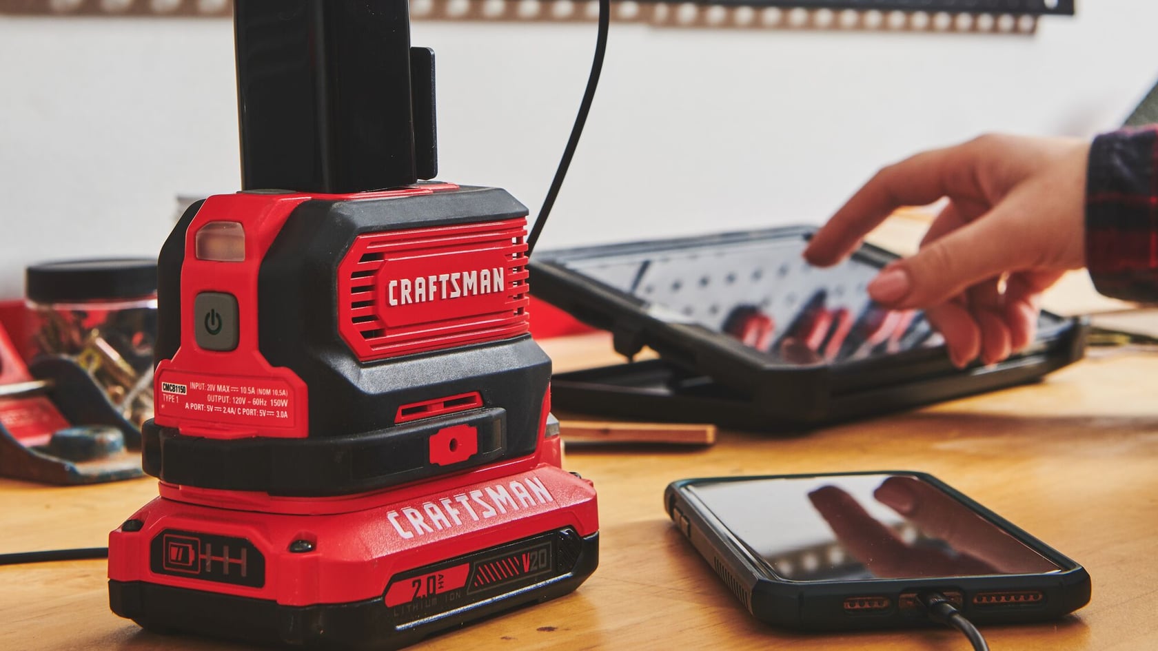 CRAFTSMAN (R) V20 Portable Power Station/inverter (150 Watt) (CMCB1150B) on a workbench, charging a phone and tablet.
